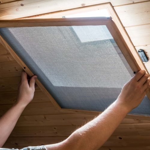 Unrecognizable person is installing a wooden homemade mosquito net attached with magnets, front of an opened velux window on ceiling, in home interior in attic, to prevent flying insects from entering.
